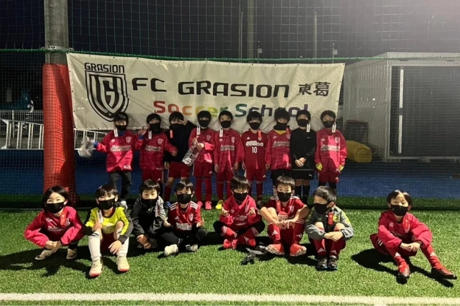 赤いサッカーユニフォームとマスクを着用した子供たちが、夜間の人工芝グラウンドで集合写真を撮っている。