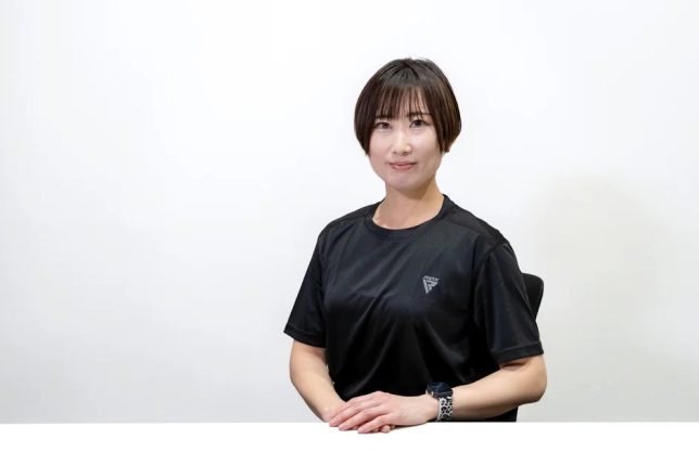 白い背景の前で、黒いスポーツTシャツを着たショートヘアの女性が座っています。