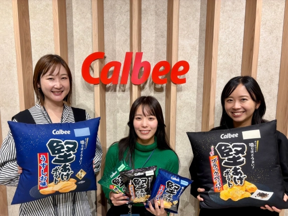 カルビーのロゴを背景に、3人の女性が笑顔でポテトチップス「堅あげポテト」の商品や、そのパッケージを模したクッションを持って立っている