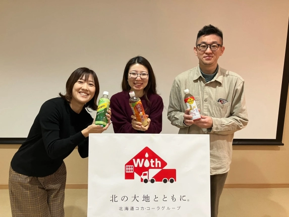 北海道コカ・コーラグループのバナーの前で、3人の従業員が笑顔で同社の飲料製品を持っている集合写真