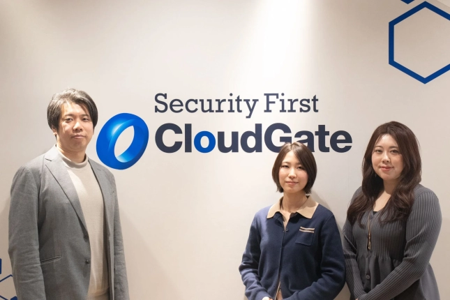 Security First CloudGateのロゴが描かれた白い壁の前で、男性1名と女性2名が並んで立っている