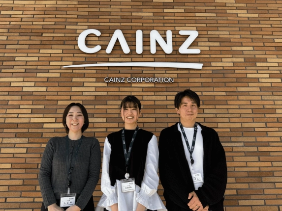 カインズのロゴが入ったレンガの壁の前で、3人の社員が笑顔で立っている集合写真