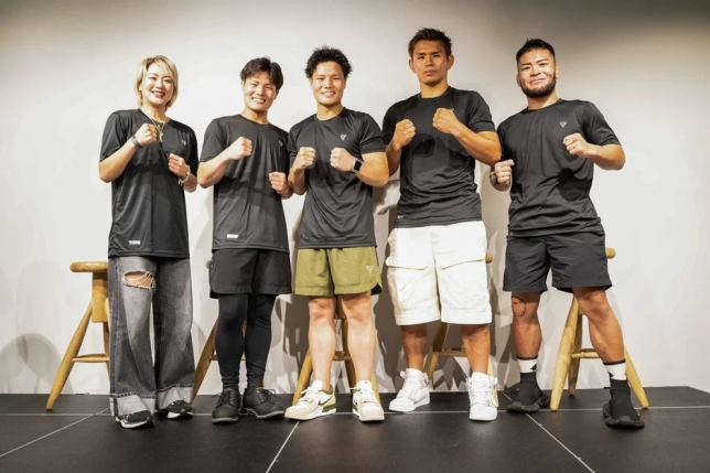 黒いトレーニングウェアを着用した5人の男女が、拳を握り笑顔でポーズをとっている集合写真です。