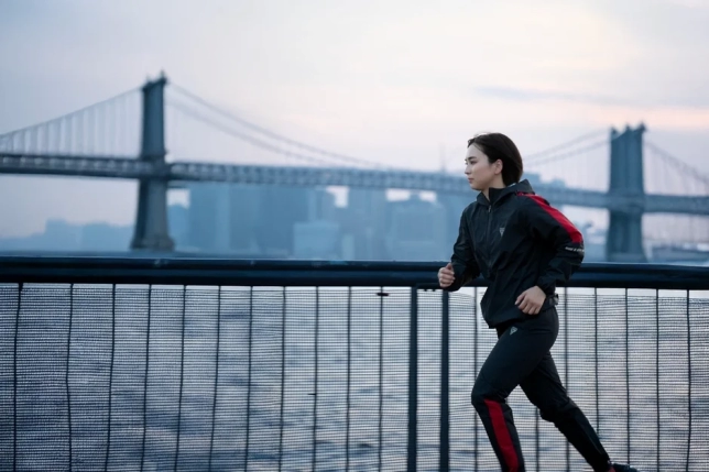 女性がスポーツウェアを着て、大きな吊り橋を背景に水辺をランニングしている様子。