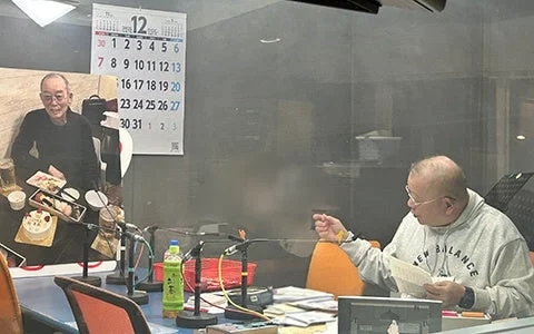 ラジオスタジオでマイクに向かう笑福亭鶴瓶さんと、新野新さんの写真パネル