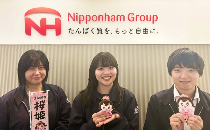 日本ハムグループのロゴ前で、3人の従業員が「桜姫」ブランドの旗やマスコットを持って笑顔でポーズ