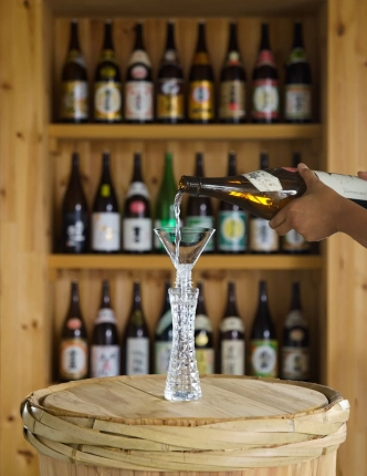 木製の棚にずらりと並んだ日本酒の瓶を背景に、手がボトルから透明なガラスの器に日本酒を注いでいる様子。木製の樽の上で行われており、伝統的な酒蔵のような雰囲気を醸し出しています。